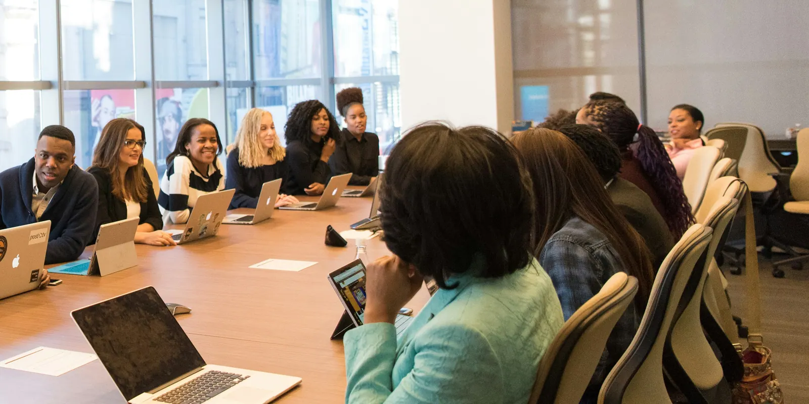 A group seated along a long conference table with laptops open, listening to the person at its head
